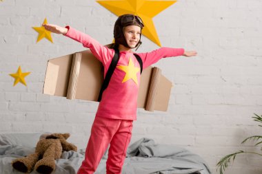 Inspired child with cardboard wings gesturing in bedroom clipart
