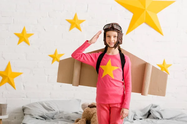 Adorable kid with cardboard wings saluting with smile