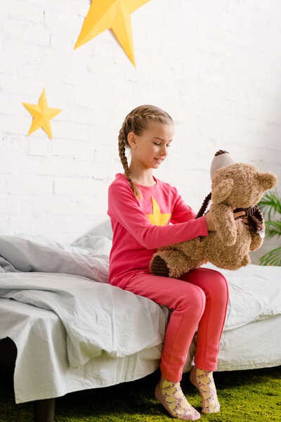 Cute kid with braids sitting on bed and looking at teddy bear
