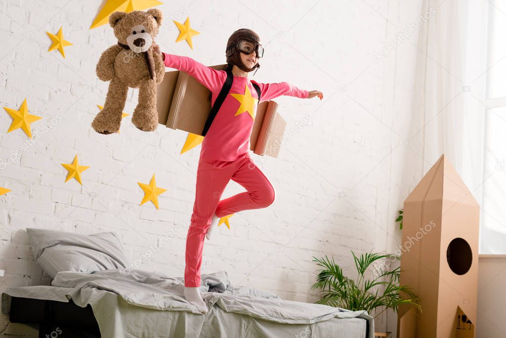 Kid with teddy bear standing on bed on one leg