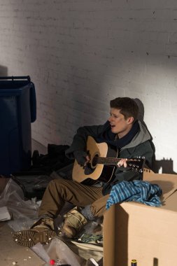 çöp ve gitar çalmak çevrili karton üzerine oturan evsiz adam