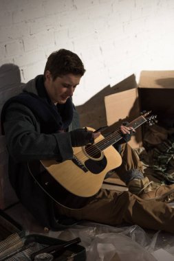 gitar tutarak ve çöp dökümü üzerinde otururken smartphone kullanarak evsiz adam
