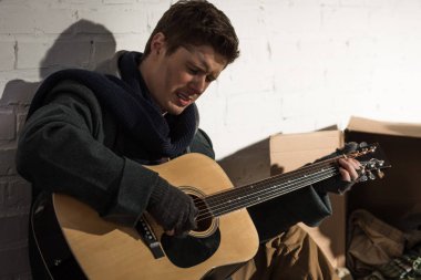 Beyaz tuğla duvar dibinde otururken gitar çalmak üzgün evsiz adam