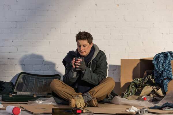 poor homeless man drinking from paper cup while sitting on cardboard surrounded by messed rubbish