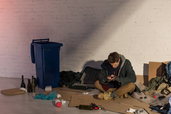 homeless man using smartphone while sitting on cardboard surrounded by rubbish dump