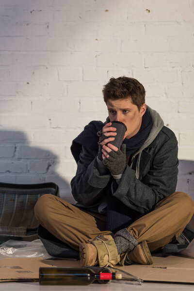 sad homeless man drinking from paper cup while sitting on cardboard by brick wall