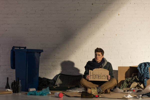 depressed homeless man sitting on cardboard surrounded by garbage and holding cardboard card with "homeless" handwritten lettering