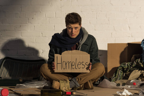 sad man sitting on rubbish dump and holding cardboard card with "homeless" handwritten text