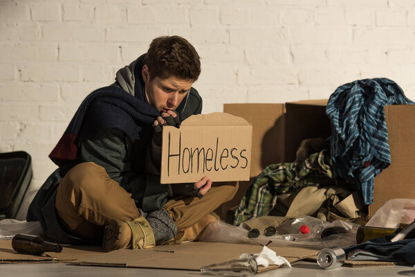 misery man sitting on garbage dump and holding piece of cardboard with "homeless" inscription