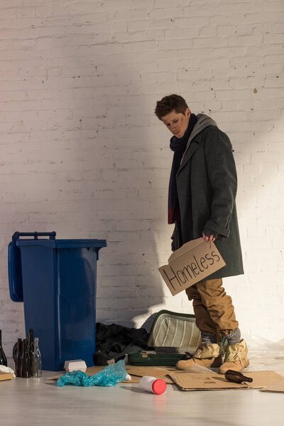 homeless misery man standing near trash container and holding cardboard card with "homeless" handwritten text