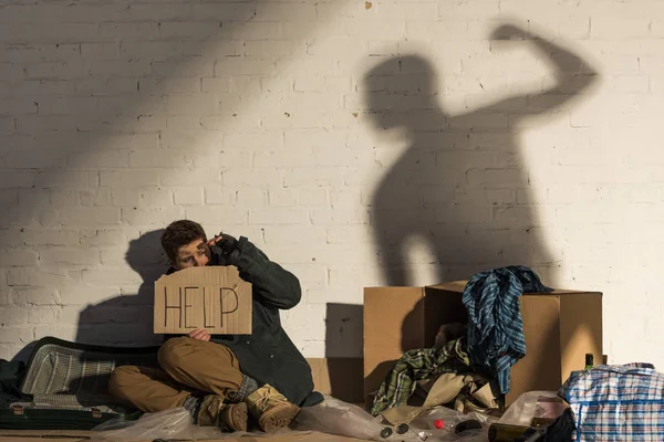 受惊的无家可归的人拿着卡片与 的字母 而坐在砖墙与人的影子举起手用拳头 — 图库照片