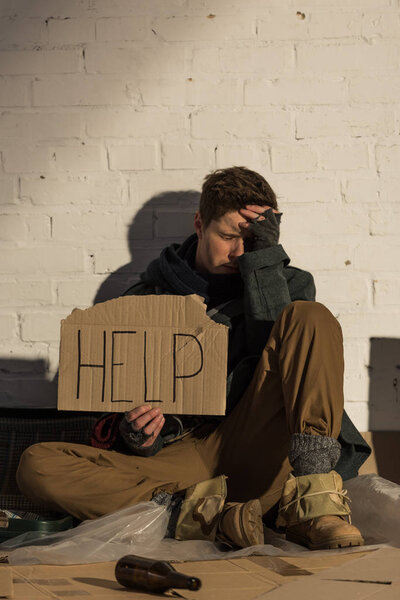 отчаявшийся бездомный сидит у кирпичной стены и держит кусок картона с надписью "помощь"
