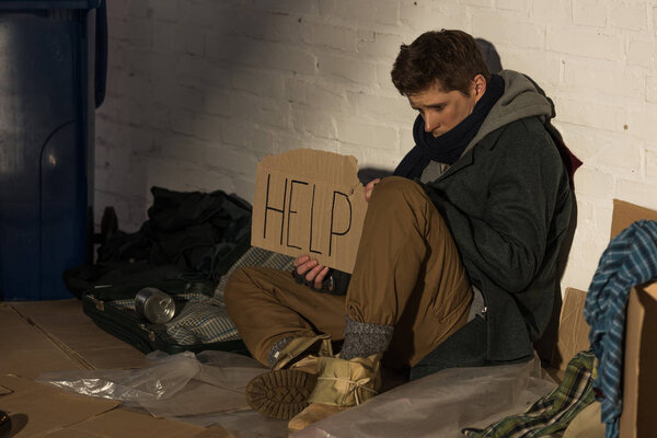 depressed homeless man holding piece of cardboard with "help" inscription while sitting on trash dump