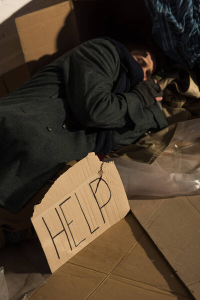 exhausted homeless man with "help" inscription card sleeping on cardboard