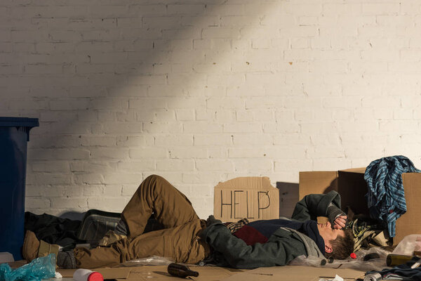 homeless man lying on cardboard surrounded by rubbish under white brick wall
