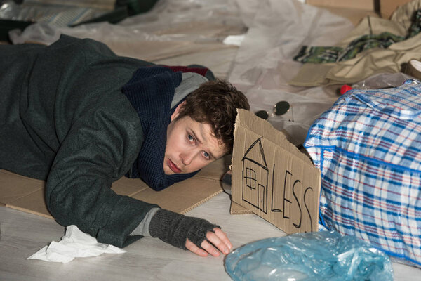 despaired homeless man lying on cardboard in rubbish dump, with symbol of house and "less" inscription on card
