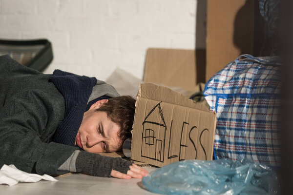 poor homeless man sleeping on rubbish damp with symbol of house and "less" lettering on piece of cardboard 