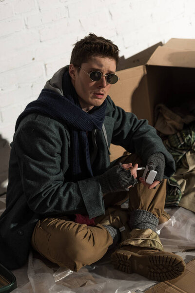 homeless man sitting by white brick wall and holding cigarette pack