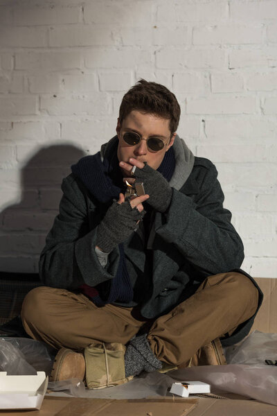 homeless man lighting cigarette while sitting by brick wall