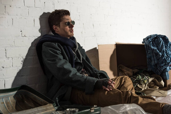 homeless man smoking while sitting by white brick wall