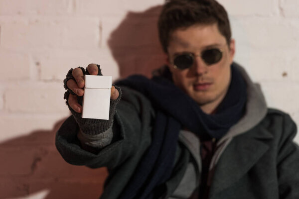 selective focus of homeless man in fingerless gloves and dark glasses holding cigarette pack in stretched hand