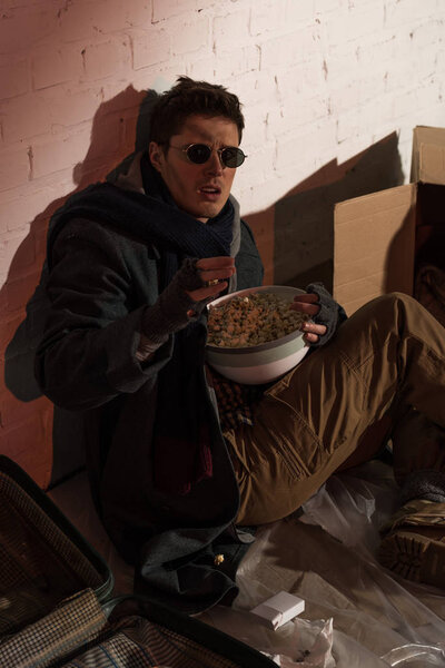 homeless man sitting by brick wall and holding bowl of popcorn 