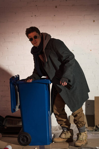homeless man in dark glasses rummaging in blue trash container