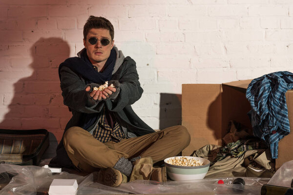 homeless man sitting by white brick wall and stretching hands with popcorn