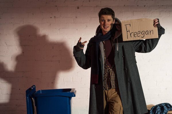 homeless man holding cardboard card with "freegan" lettering and showing victory sign