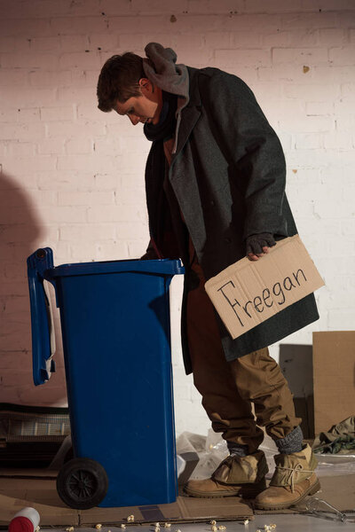 homeless man rummaging in rubbish container while holding cardboard card with "freegan" handwritten lettering