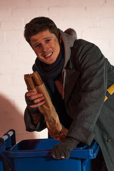 счастливый бездомный держит хлеб багет, стоя возле мусорного контейнера
