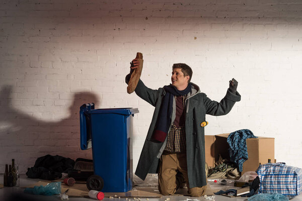 homeless man standing on knees and holding bread baguette in raised hand