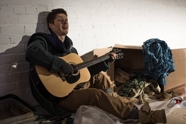 Бездомный играет на гитаре и поет, сидя на помойке.
