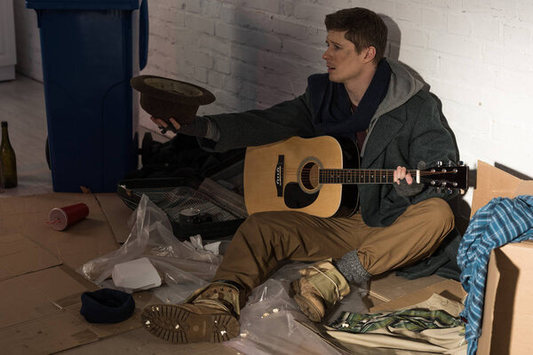 homeless man with guitar sitting on rubbish dump and holding hat in stretched hand