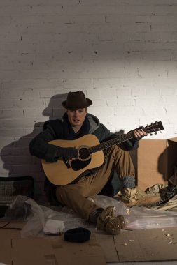 Evsiz karanlık giysili adam ve çöp dökümü üzerinde oturan ve gitar çalmak şapka