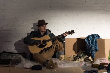 homeless man sitting playing guitar while sitting on cardboard surrounded by rubbish  clipart