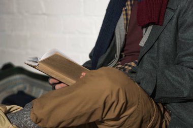 cropped view of homeless man reading book while sitting by white brick wall clipart