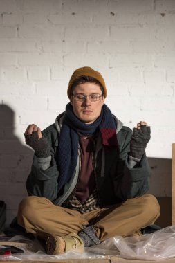 homeless man sitting in lotus pose with closed eyes near white brick wall clipart