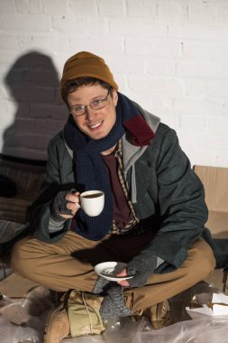 smiling homeless man drinking coffee while sitting by white brick wall surrounded by rubbish clipart