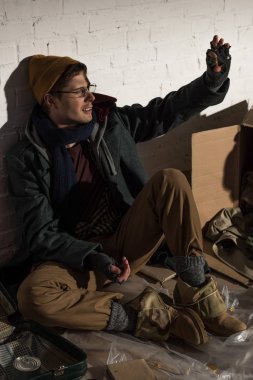 homeless beggar man sitting by brick wall and waving hand clipart