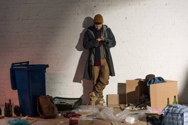 evsiz adam çöp tarafından çevrili beyaz tuğla duvar tarafından duran Smartphone kullanarak