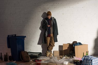 homeless man using smartphone while standing on rubbish dump clipart
