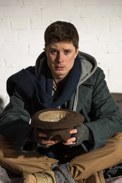 Begging Homeless Man Holding Hat While Sitting White Brick Wall — Stock Photo, Image