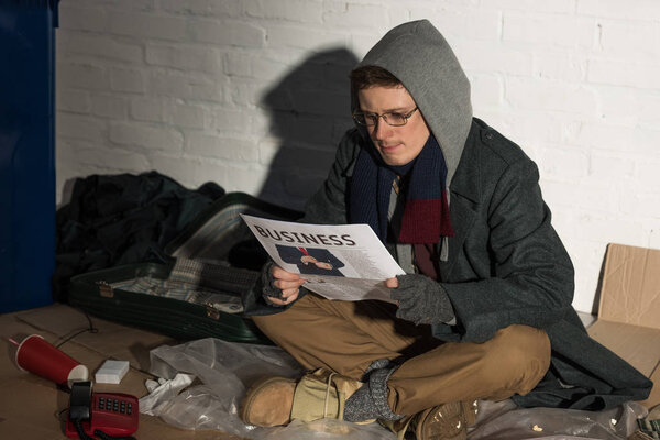 homeless man reading business newspaper while sitting on rubbish dump