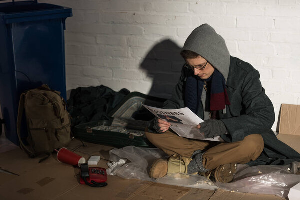 homeless man in hood and glasses reading business newspaper while sitting by brick wall