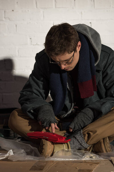 Бездомный в очках с помощью винтажного телефона сидит у кирпичной стены
