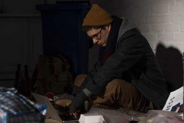 Бездомный использует ноутбук, сидя на свалке.
