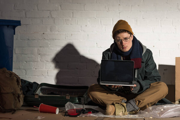 серьезный бездомный с ноутбуком, сидя у кирпичной стены на мусорной свалке
