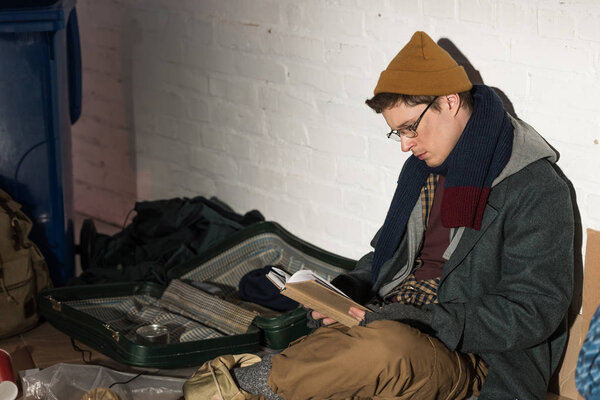 homeless man in glasses reading book while sitting on rubbish dump