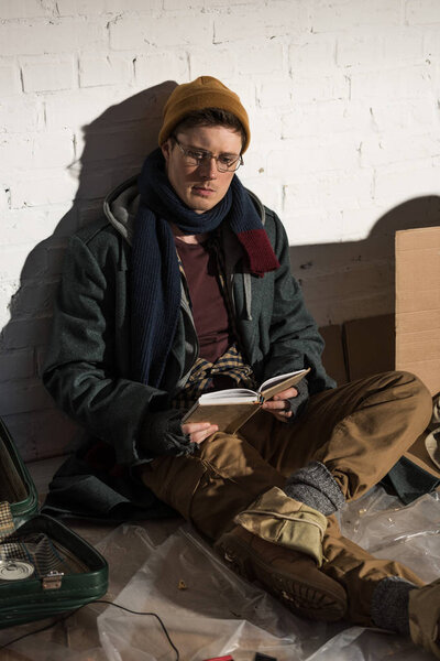 homeless man reading book while sitting by white brick wall 